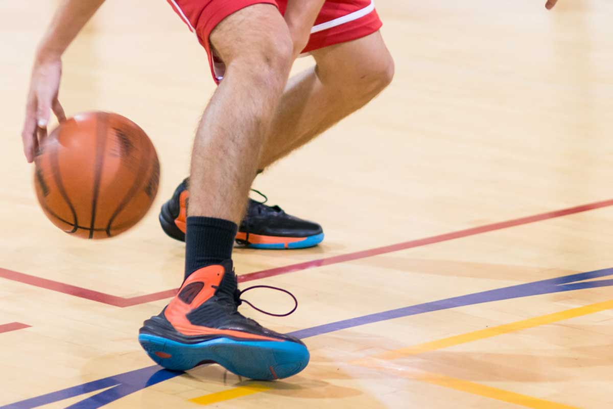 A high school basketball player dribbles between his legs with his basketball shoes grips on the floor, How To Add Grip To Basketball Shoes [5 Crucial Tips]