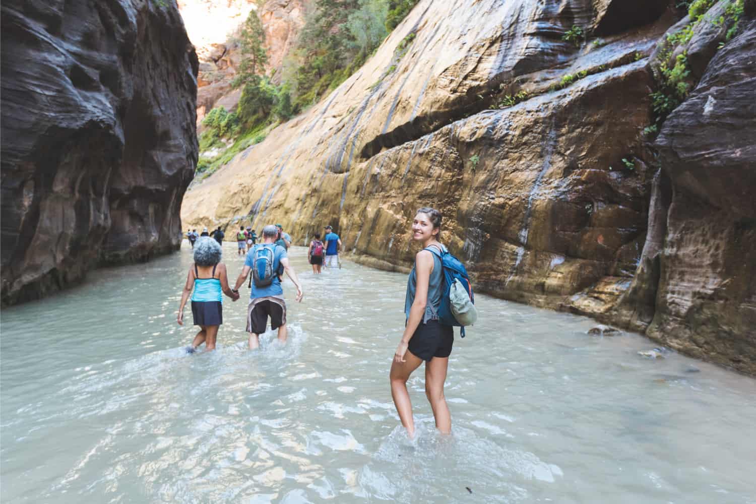 Different group of hikers wade through knee-deep water in a beautiful red slot canyon