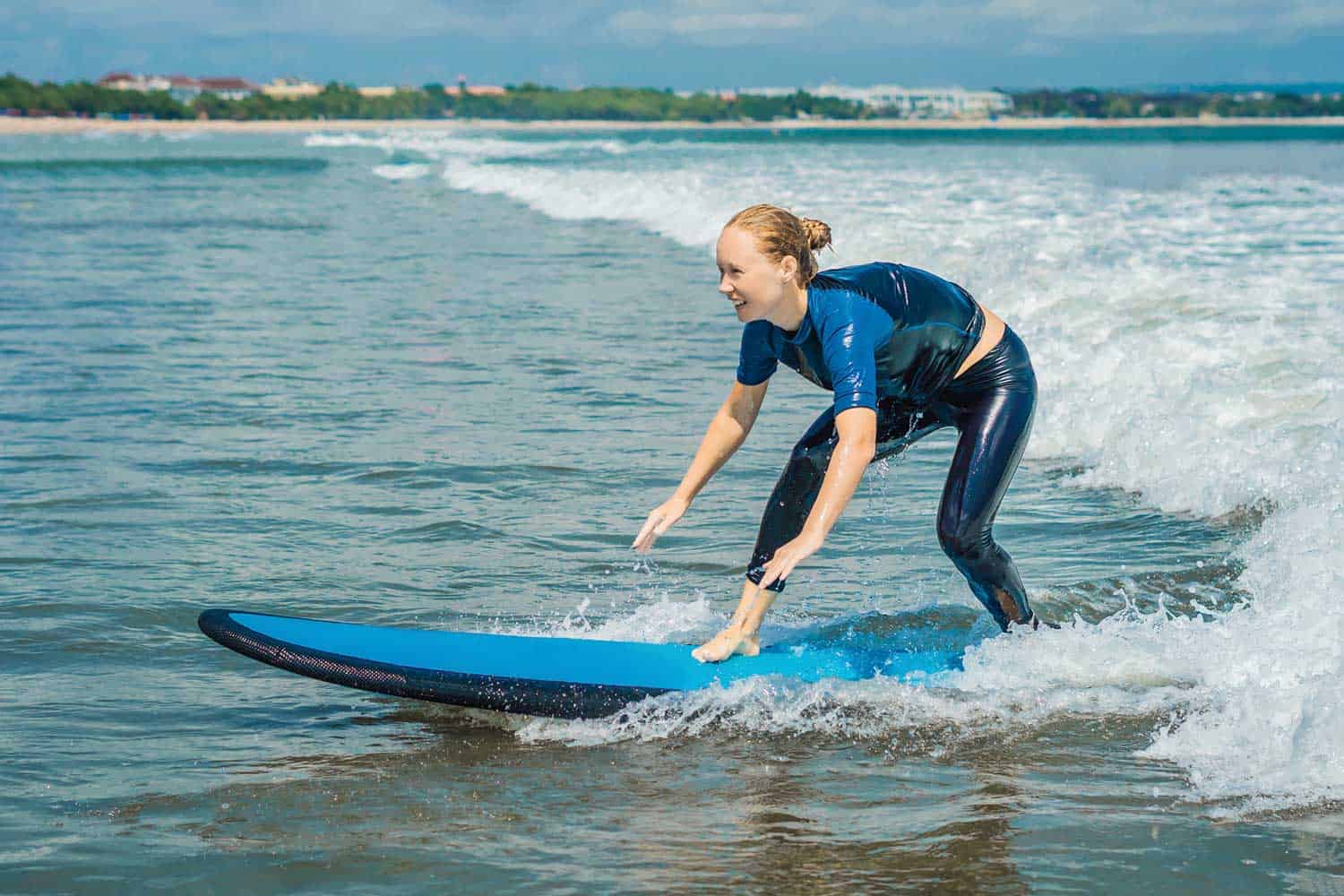 Joyful young woman beginner surfer with blue surfboard having fun on small waves