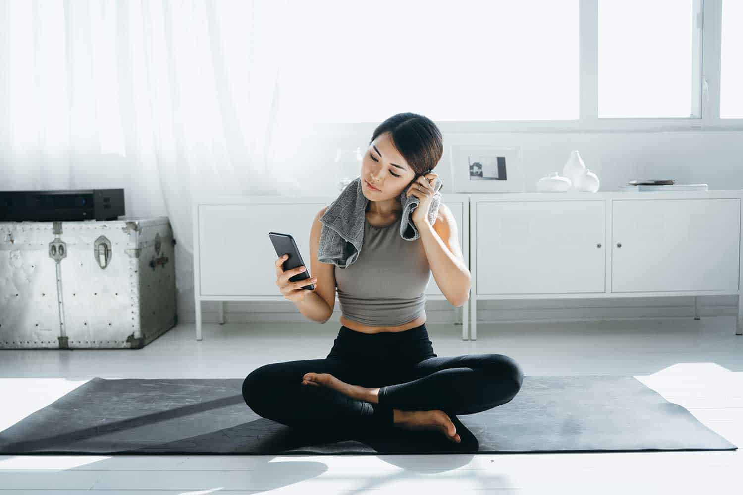 Young asian sports woman wiping sweat with a towel around her neck and using smartphone after exercising at home in the morning