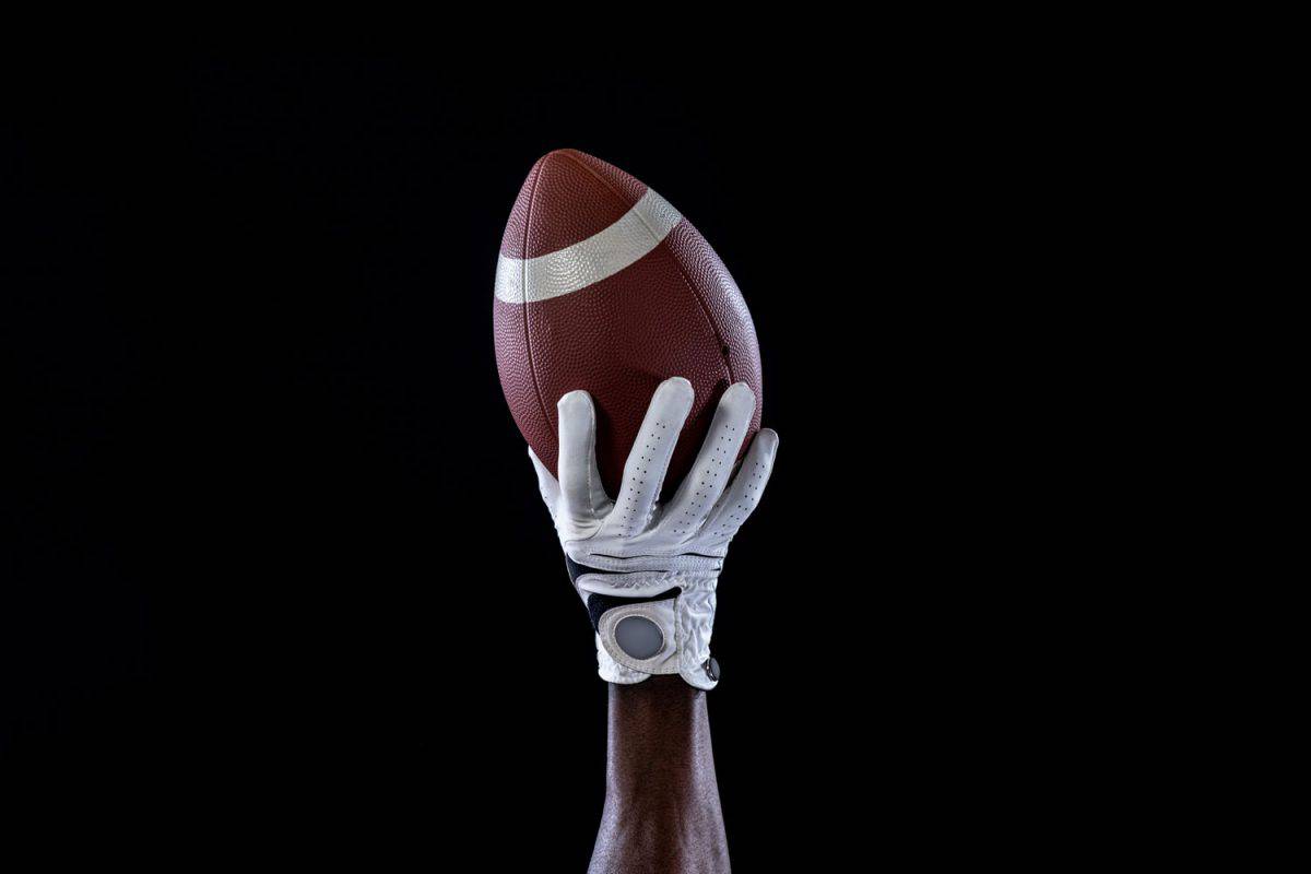 A football player wearing white football gloves and holding a football on a dark background