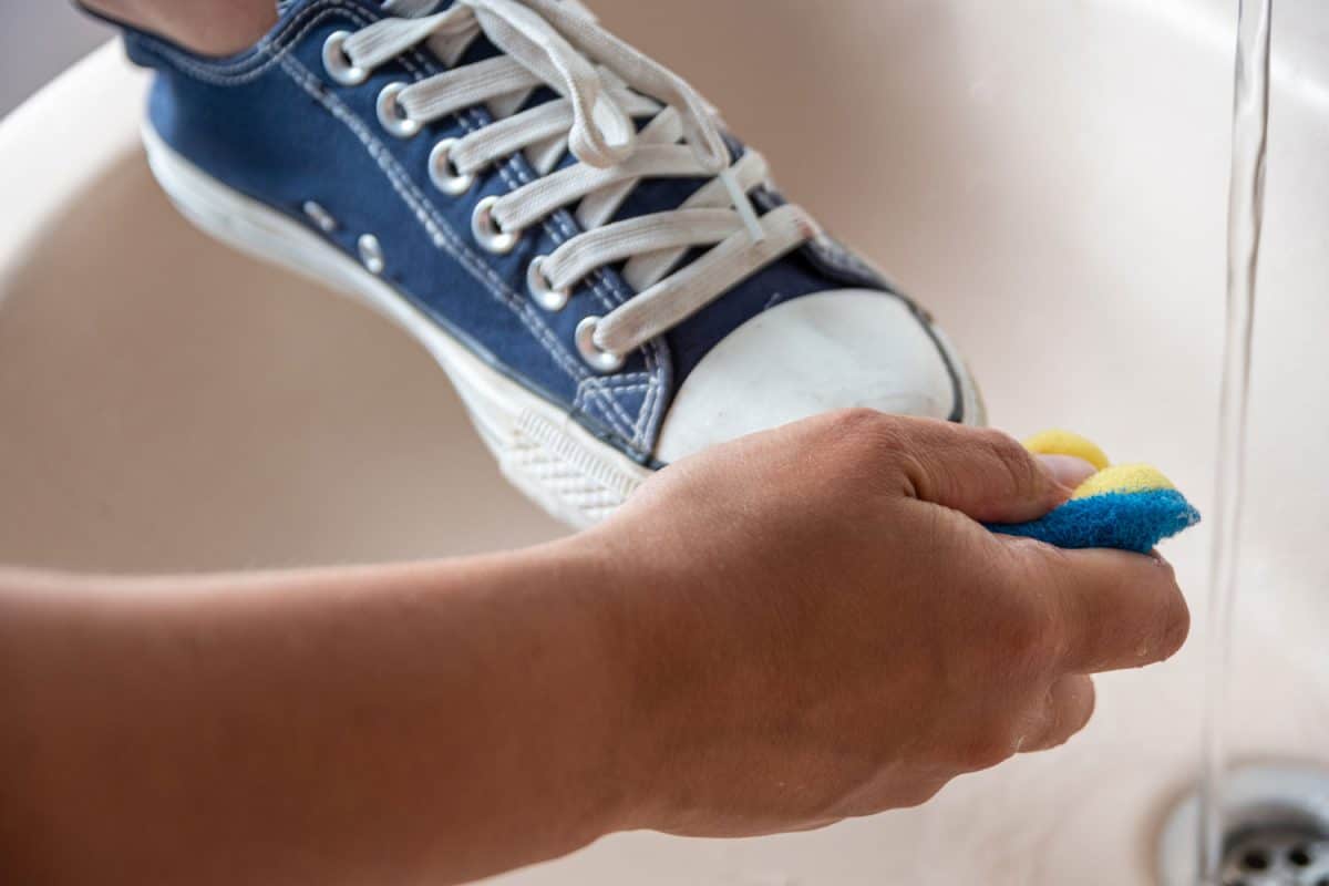 Adult is cleaning a blue laced sneaker in sink unsing a blue yellow sponge